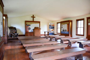 A room inside the Lac qui Parle Mission filled with wooden benches facing a podium at the front of the room. 