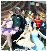 Cast posing for a picture. Some wearing ballerina costumes, another in a blue dress, a man in an old fashioned suit. 